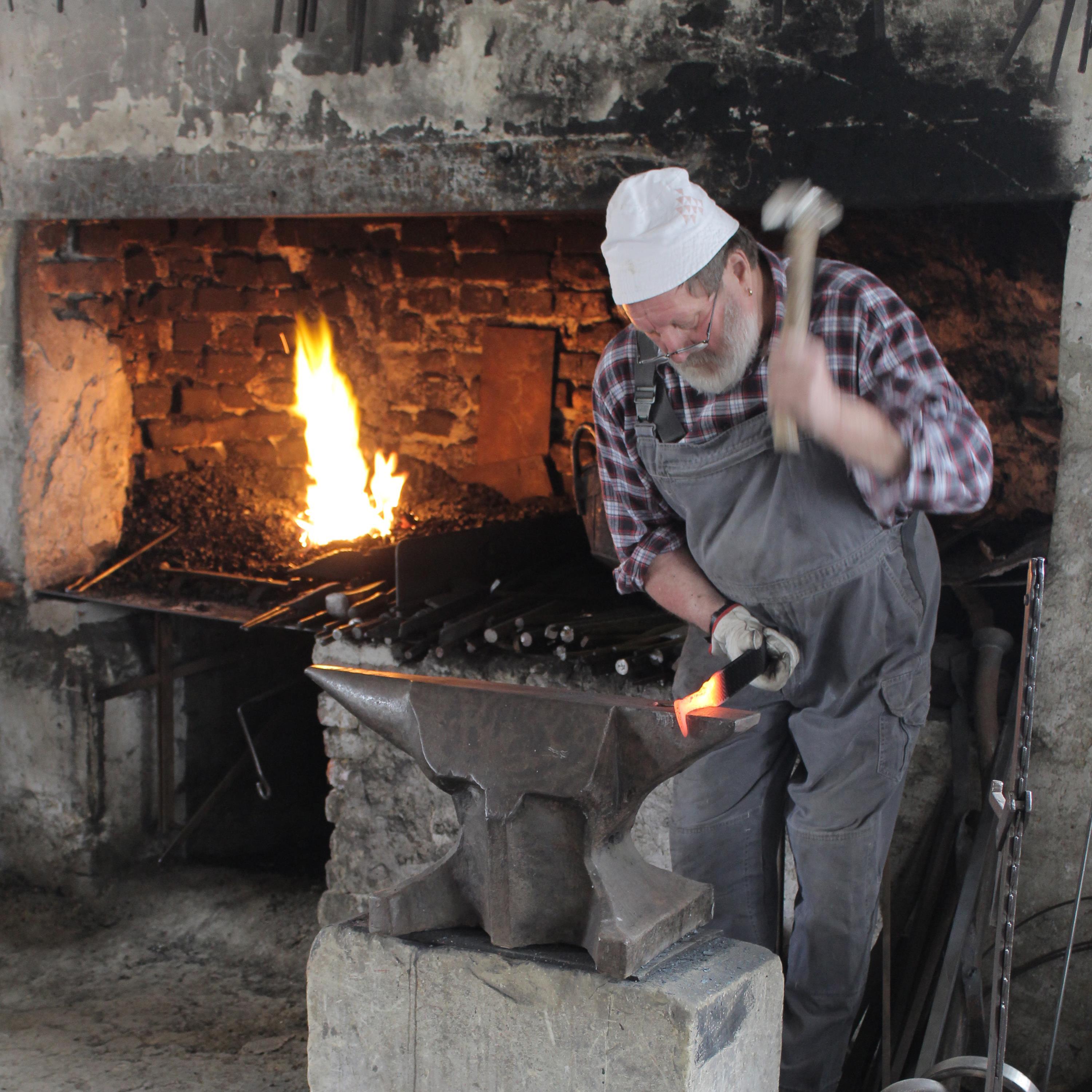 Schmied formt Metallstück am Amboss, im Hintergrund lodert Feuer